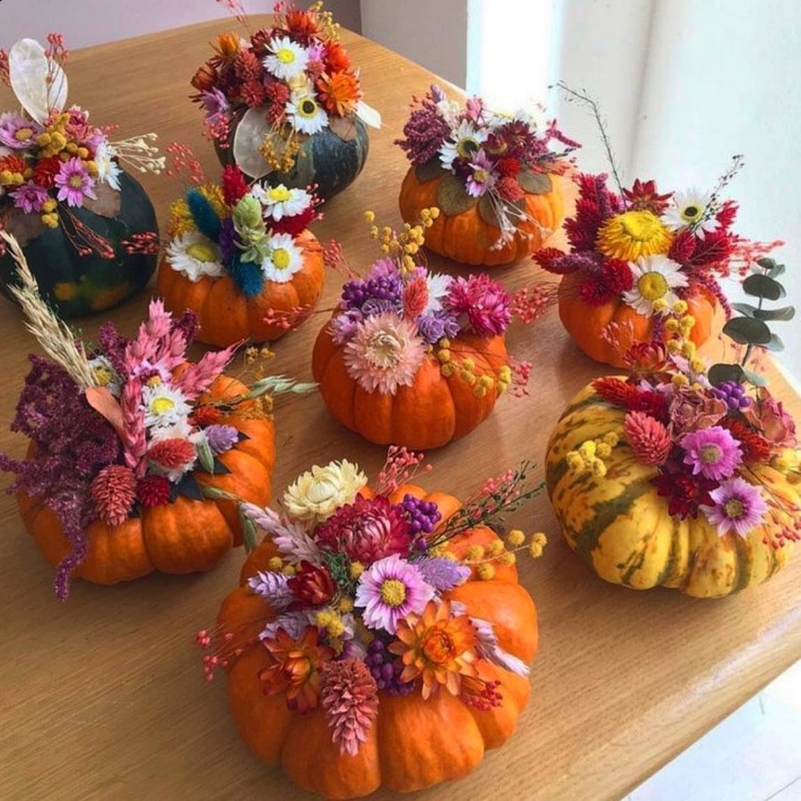 Small pumpkins filled with colorful autumn flowers arranged on a light wooden table.