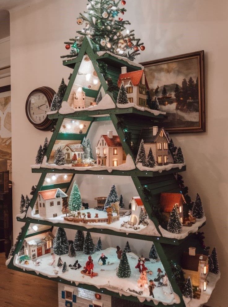 Green wooden Christmas tree shelf decorated with miniature snowy village houses and fairy lights.