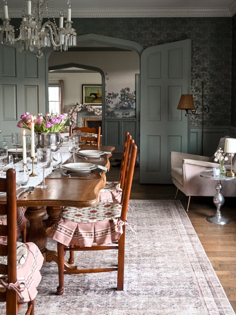 Wooden dining table with pink cushions in a dark grey paneled room with chandelier.