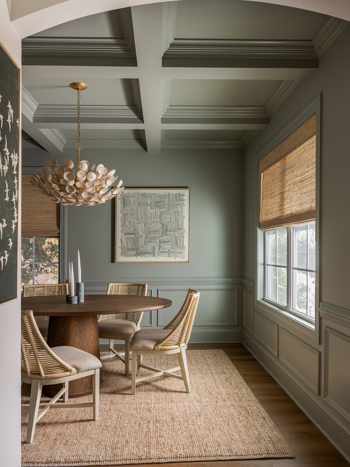 Sage green dining room with coffered ceiling, wooden table, and woven chairs.